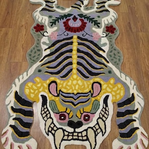 May include: A decorative tiger-shaped rug with a whimsical design. The rug features a light purple background with black stripes, yellow accents, and floral details. The rug has a white border and is placed on a wooden floor.