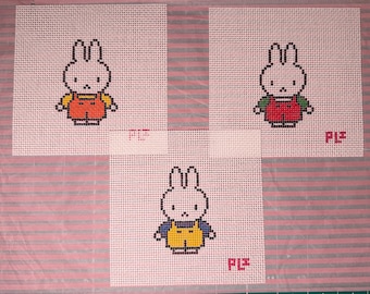 May include: Three square white plastic canvas sheets featuring cross-stitch patterns of a rabbit character. The top left rabbit wears orange overalls, the top right wears red overalls with green sleeves, and the bottom rabbit wears yellow overalls.