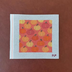 May include: A square needlepoint canvas featuring a repeating pattern of small pumpkins in shades of yellow, orange, and red against a coral background. The canvas has a white border and a single orange vertical line on the left side with PLZ text.