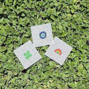 May include: Three square white plastic canvas sheets featuring needlepoint designs. One sheet displays a green four-leaf clover, another shows a blue and white evil eye symbol, and the third features a small multicolored rainbow arc design.