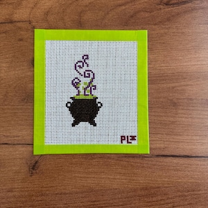 May include: A square needlepoint canvas featuring a black cauldron with bright green bubbling liquid and purple swirling steam. The design is bordered by a lime green tape frame and includes the stitched initials PL in the bottom right corner.