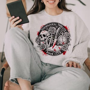 May include: A light gray sweatshirt featuring a circular graphic with a skull, roses, a knife, and a pocket watch. The text "PSYCHOLOGICAL THRILLER" and "BOOK CLUB" are printed around the design. The design has red blood splatter accents.