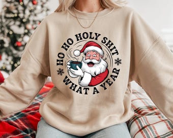 May include: Beige sweatshirt featuring a graphic of Santa Claus holding a mug, with the text "HO HO HOLY SHIT WHAT A YEAR" in a circular design. Santa is wearing a red hat and coat. The sweatshirt is a neutral color.