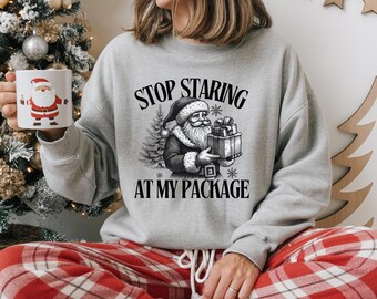 May include: A light gray sweatshirt with the text "STOP STARING AT MY PACKAGE" and a black and white Santa Claus graphic holding a gift. The person is also holding a mug with a Santa Claus design. The person is wearing red and white plaid pajama pants.