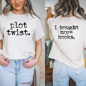 May include: Cream-colored t-shirt with the text "plot twist." on the front and "i bought more books." on the back in black, typewriter-style font. The shirt is paired with light blue jeans.