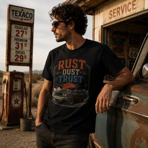 May include: A black short-sleeved t-shirt featuring a graphic print of a vintage car in a desert landscape with a setting sun. The text "RUST DUST TRUST" is printed in orange and blue above the car illustration. The shirt has a crew neckline.