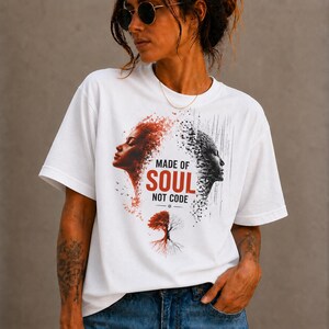 Puede incluir: Una camiseta blanca de manga corta con un estampado gráfico de dos perfiles que miran en direcciones opuestas. El lado izquierdo es rojo con elementos orgánicos, mientras que el derecho es negro con patrones de píxeles digitales. Texto: MADE OF SOUL NOT CODE.