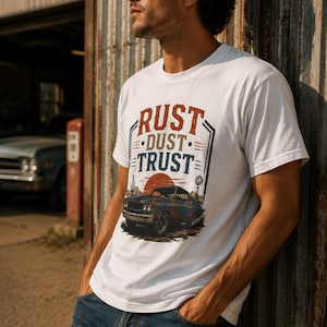 May include: A white short-sleeve t-shirt featuring a graphic print with the text RUST DUST TRUST in red, tan, and blue. The design includes a vintage car, a setting sun, a cactus, and a windmill, all rendered in a distressed, weathered style.