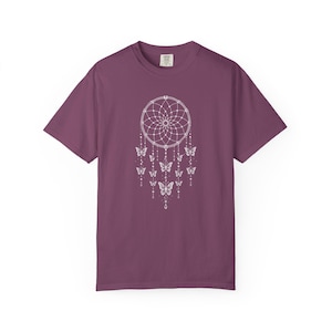 May include: A plum-coloured short-sleeve t-shirt featuring a white graphic print on the chest. The design depicts a circular dreamcatcher web with multiple dangling strings, beads, and several butterflies of varying sizes integrated into the pattern.