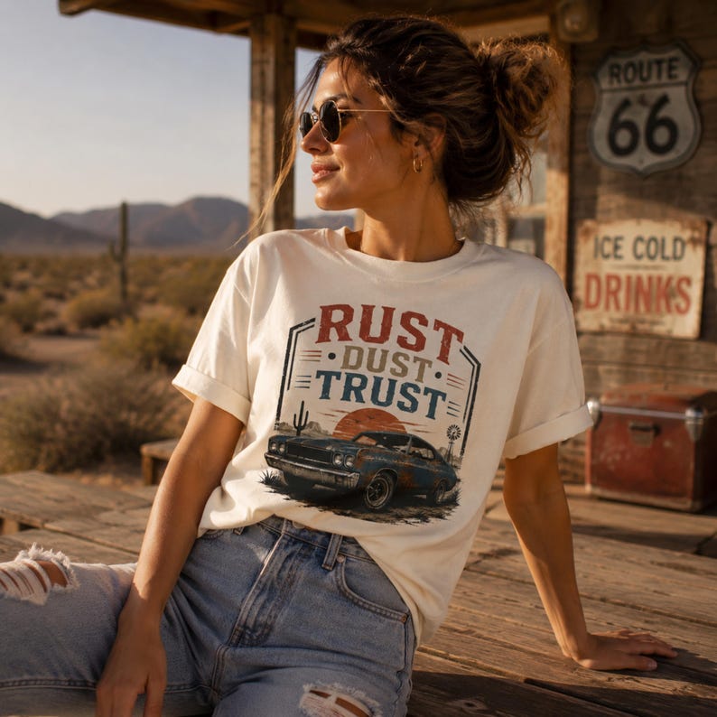 May include: Cream-colored t-shirt with the text "RUST • DUST • TRUST" above a graphic of a classic car at sunset. The shirt is paired with blue jeans. The scene is set outdoors with desert scenery.