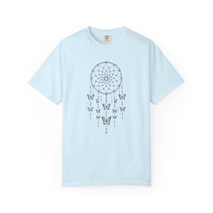 May include: A light blue short-sleeved t-shirt featuring a black line art graphic of a dreamcatcher centred on the chest. The dreamcatcher design includes intricate web patterns, hanging beaded strings, and several butterfly illustrations.