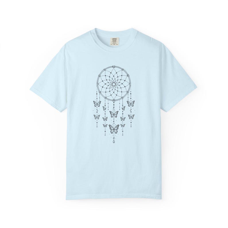 May include: A light blue short-sleeved t-shirt featuring a black line art graphic of a dreamcatcher centred on the chest. The dreamcatcher design includes intricate web patterns, hanging beaded strings, and several butterfly illustrations.