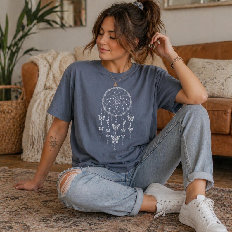 May include: A dusty blue t-shirt featuring a white dreamcatcher design with butterflies. The shirt is a relaxed fit and has short sleeves. The model is wearing light wash jeans and white sneakers. The background includes a brown leather couch and a woven basket.