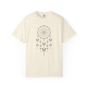 May include: A cream-coloured short-sleeve t-shirt featuring a black line-art graphic of a dreamcatcher centred on the chest. The dreamcatcher design includes hanging strings adorned with small beads and several butterfly illustrations.