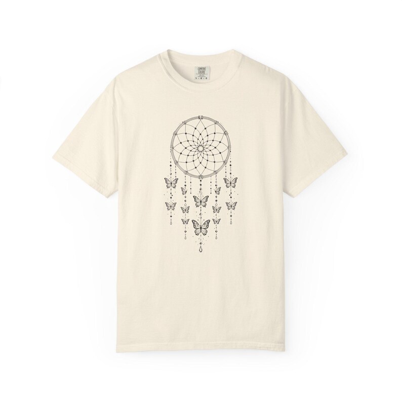 May include: A cream-coloured short-sleeve t-shirt featuring a black line-art graphic of a dreamcatcher centred on the chest. The dreamcatcher design includes hanging strings adorned with small beads and several butterfly illustrations.