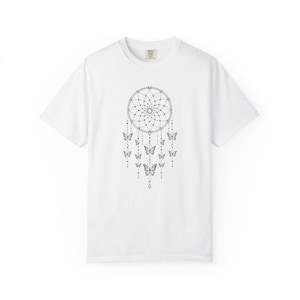 May include: A white short-sleeved t-shirt featuring a black line art graphic on the chest. The design depicts a circular dreamcatcher web with multiple dangling strings adorned with small beads and several butterflies of varying sizes.