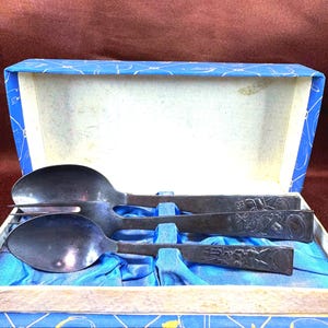 May include: A set of three dark metal utensils consisting of two spoons and one fork, each featuring engraved designs on the handles. The utensils are presented in a rectangular blue box with a white interior and blue fabric lining.