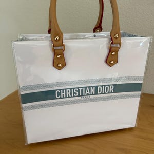 May include: White tote bag with clear sides and brown handles. The bag features the text "CHRISTIAN DIOR PARIS" in black on a silver and teal stripe across the front. The handles are tan with red trim.