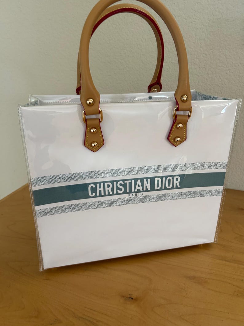 May include: White tote bag with clear sides and brown handles. The bag features the text "CHRISTIAN DIOR PARIS" in black on a silver and teal stripe across the front. The handles are tan with red trim.