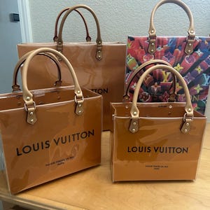 May include: Four Louis Vuitton tote bags in varying sizes. The bags are a glossy, caramel-colored material with tan handles and gold-tone hardware. The largest bag has a solid color, while another features a colorful print. Each bag has the brand name printed on the front.