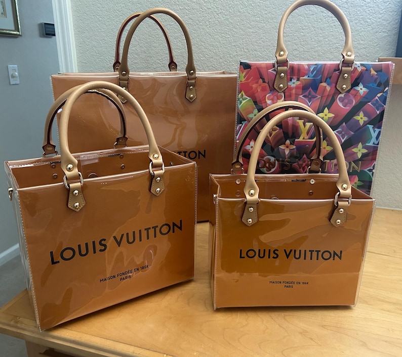 May include: Four Louis Vuitton tote bags in varying sizes. The bags are a glossy, caramel-colored material with tan handles and gold-tone hardware. The largest bag has a solid color, while another features a colorful print. Each bag has the brand name printed on the front.
