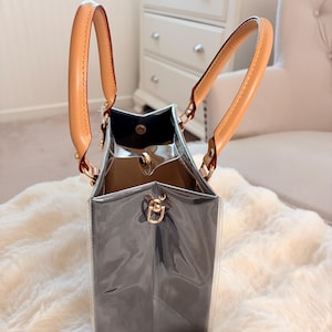 May include: A clear tote bag with a dark gray interior and tan leather handles. The bag has gold-colored hardware and a rectangular shape. The handles are attached with gold-colored metal rings.