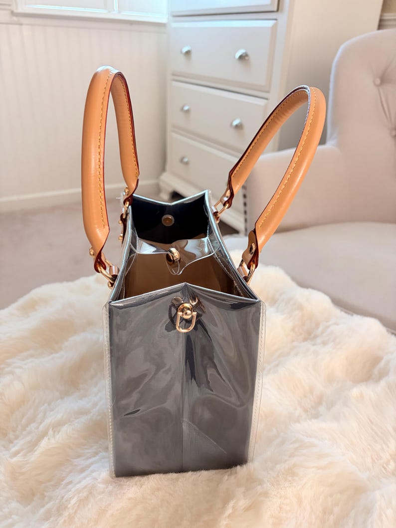 May include: A clear tote bag with a dark gray interior and tan leather handles. The bag has gold-colored hardware and a rectangular shape. The handles are attached with gold-colored metal rings.