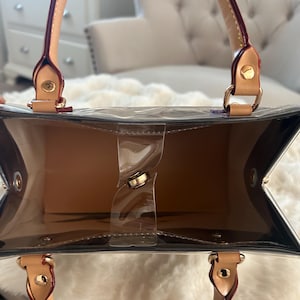 May include: A clear tote bag with tan interior and handles. The bag has a rectangular shape with a clear plastic body and tan leather handles. The handles are attached with gold-colored hardware. The bag is empty.