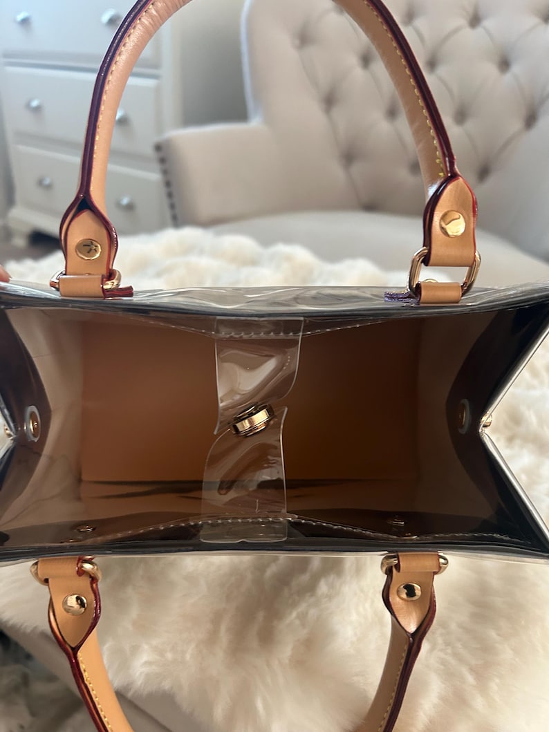 May include: A clear tote bag with tan interior and handles. The bag has a rectangular shape with a clear plastic body and tan leather handles. The handles are attached with gold-colored hardware. The bag is empty.