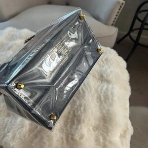 May include: A navy blue handbag with gold-colored hardware, encased in clear plastic. The bag has a textured appearance and is rectangular in shape. The corners of the bag have gold-colored studs. The bag is sitting on a white, fluffy surface.