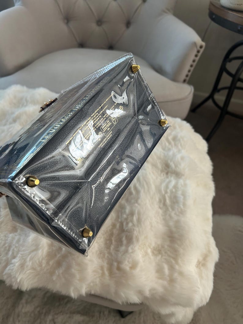 May include: A navy blue handbag with gold-colored hardware, encased in clear plastic. The bag has a textured appearance and is rectangular in shape. The corners of the bag have gold-colored studs. The bag is sitting on a white, fluffy surface.