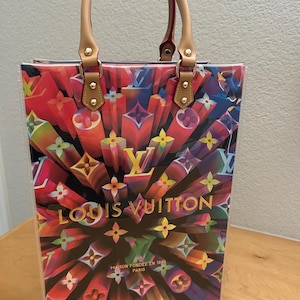 May include: A colorful tote bag with a vibrant, abstract design featuring the Louis Vuitton logo in various colors. The bag has tan leather handles and accents, with gold-colored hardware. The text "LOUIS VUITTON" is prominently displayed.
