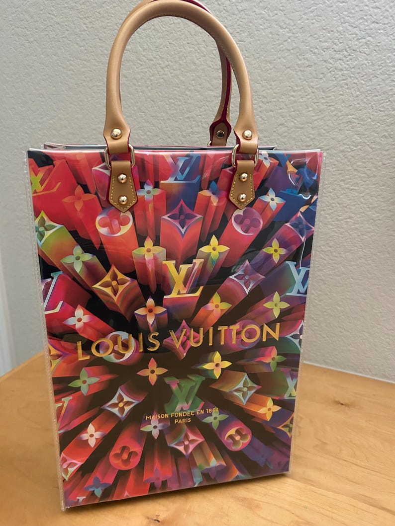 May include: A colorful tote bag with a vibrant, abstract design featuring the Louis Vuitton logo in various colors. The bag has tan leather handles and accents, with gold-colored hardware. The text "LOUIS VUITTON" is prominently displayed.