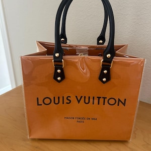 May include: A clear tote bag with a brown interior and black handles. The bag features the "LOUIS VUITTON" logo in black, along with "MAISON FOND&Eacute;E EN 1854 PARIS". The handles are attached with gold-colored hardware.