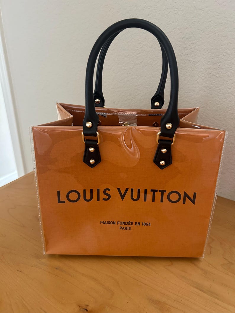 May include: A clear tote bag with a brown interior and black handles. The bag features the "LOUIS VUITTON" logo in black, along with "MAISON FOND&Eacute;E EN 1854 PARIS". The handles are attached with gold-colored hardware.