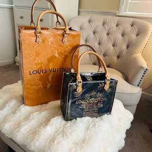May include: Two Louis Vuitton handbags. The larger bag is a glossy orange with tan handles and the brand name printed on the front. The smaller bag is black with gold accents and the Louis Vuitton logo. Both bags have tan handles.