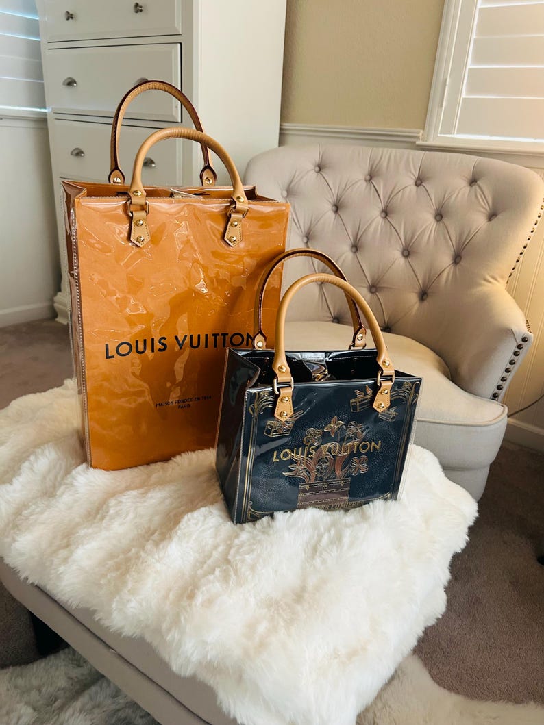 May include: Two Louis Vuitton handbags. The larger bag is a glossy orange with tan handles and the brand name printed on the front. The smaller bag is black with gold accents and the Louis Vuitton logo. Both bags have tan handles.