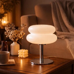 Peut inclure: Lampe de table blanche en verre à deux niveaux, avec une base chromée, posée sur une table en bois foncé. La lampe est le point central, dégageant une lumière douce. On aperçoit également une bougie, un vase avec des fleurs séchées et une tasse.