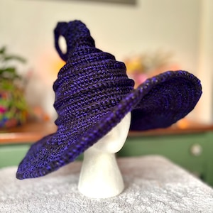 May include: A pointed hat crafted from dark purple and black yarn with a wide, curved brim. The hat features a textured, ribbed crochet pattern throughout the conical crown and the circular brim, creating a distinct two-tone color effect.