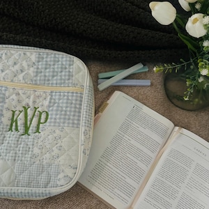 May include: A quilted fabric case featuring a light blue and white patchwork pattern with a gingham and floral design. The letters KVP are embroidered in green thread on the front. The case has a zipper closure and a light-colored piping trim.