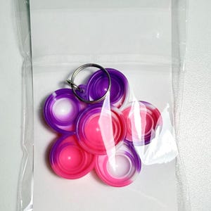 May include: A collection of colorful, circular silicone fidget toys in a clear plastic bag. The toys are in shades of pink, purple, and white, attached to a silver keyring. The design is a flower-like arrangement.