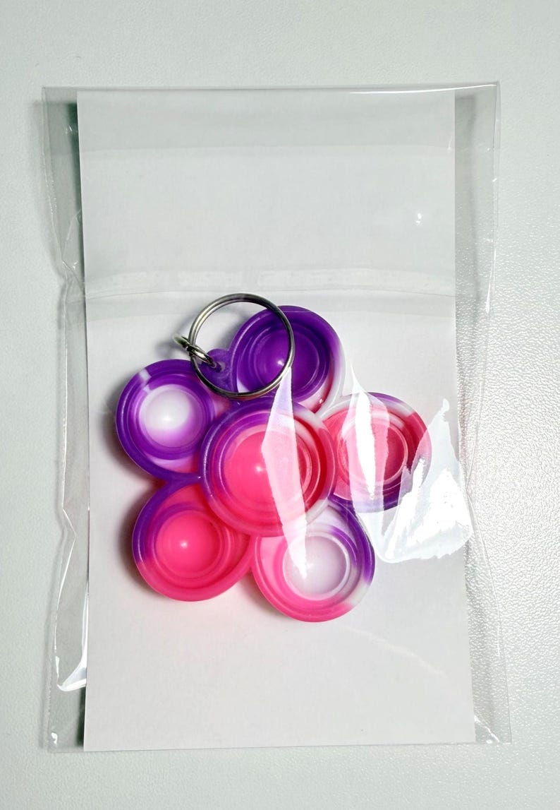 May include: A collection of colorful, circular silicone fidget toys in a clear plastic bag. The toys are in shades of pink, purple, and white, attached to a silver keyring. The design is a flower-like arrangement.