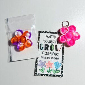 May include: Two flower-shaped silicone sensory toys and a rectangular card. One toy is orange, purple, and white in a clear bag. The other is pink and white with a metal keychain. The card reads: WATCH YOURSELF GROW THIS YEAR! LOVE, MS. EVERLY.