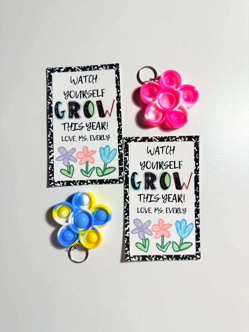 May include: Two rectangular cards with black and white speckled borders featuring the text WATCH YOURSELF GROW THIS YEAR! LOVE, MS. EVERLY and three hand-drawn flowers. Two flower-shaped silicone fidget keychains in neon pink and blue-yellow-white.