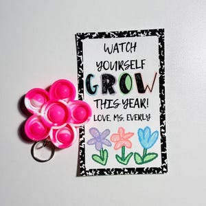 May include: A flower-shaped silicone fidget keychain in pink and white, paired with a rectangular card featuring the text WATCH YOURSELF GROW THIS YEAR! LOVE, MS. EVERLY and illustrations of three colorful flowers with a black and white border.
