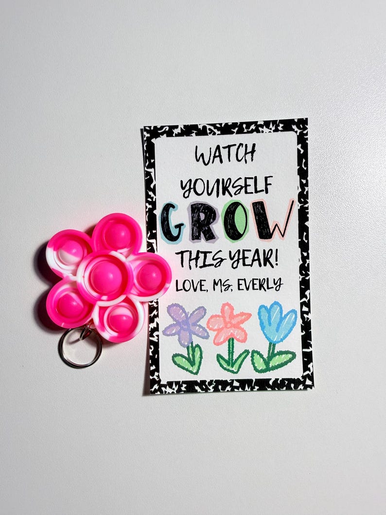 May include: A flower-shaped silicone fidget keychain in pink and white, paired with a rectangular card featuring the text WATCH YOURSELF GROW THIS YEAR! LOVE, MS. EVERLY and illustrations of three colorful flowers with a black and white border.