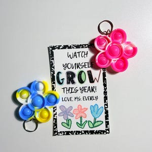 May include: Two flower-shaped silicone sensory keychains in pink-white and blue-yellow swirl patterns, accompanied by a rectangular card with black-and-white speckled borders featuring the text WATCH YOURSELF GROW THIS YEAR! LOVE, MS. EVERLY.