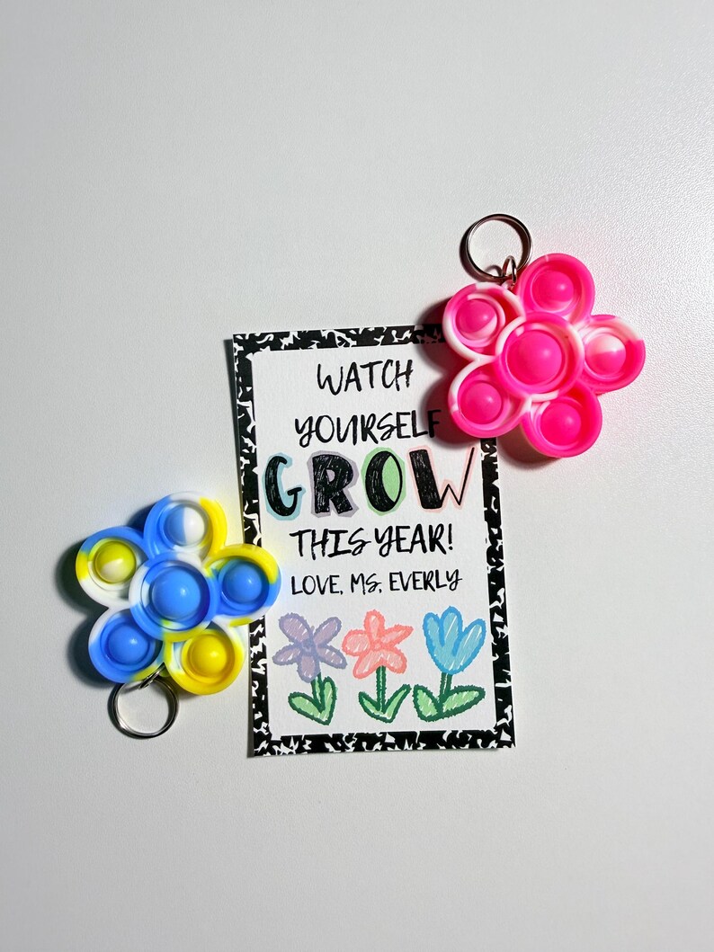 May include: Two flower-shaped silicone sensory keychains in pink-white and blue-yellow swirl patterns, accompanied by a rectangular card with black-and-white speckled borders featuring the text WATCH YOURSELF GROW THIS YEAR! LOVE, MS. EVERLY.