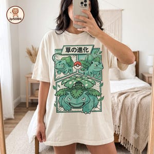 May include: An off-white short-sleeve t-shirt featuring a graphic print of three green creatures in various stages of evolution. The design includes a red and white Poké Ball icon and Japanese text that reads 草の進化, arranged in a panel layout.