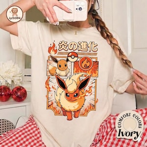 May include: An ivory t-shirt featuring a graphic print of the character Eevee evolving into Flareon. The design includes Japanese text, a Poké Ball icon, and flame motifs in orange and red. The shirt has a crew neckline and short sleeves.